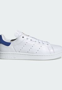 Bílé kožené tenisky s modrým akcentem na patě. Mají perforované strany, ploché tkaničky a textured bílou gumovou podrážku.