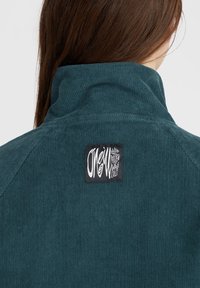 Giacca in corduroy teal con colletto alto, caratterizzata da un'etichetta nera con testo bianco sul retro. Tessuto texture e design a coste.
