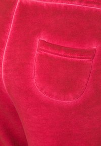 Sasietas sarkanas bikses ar mazu ieliektā aizmugurē, izgatavotas no mīksta, gluda auduma ar nedaudz spīdumu.