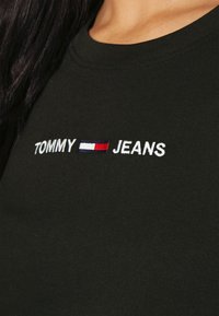 Camiseta de algodón negra con el texto bordado "TOMMY" y "JEANS" en blanco, con una barra horizontal roja y azul en el medio.
