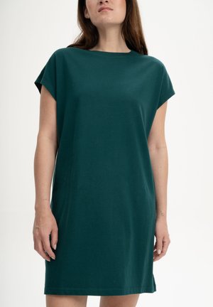 Femme portant une robe décontractée vert foncé à manches courtes et longueur genou, debout devant un fond blanc uni.
