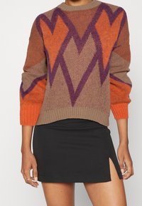 Pull en maille marron, orange et violet avec un motif géométrique en forme de cœur ; col côtelé et manches orange. Mini-jupe noire avec fente latérale.