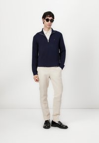 Marineblauwe trui met een ritssluiting bij de kraag, witte binnenlaag, crèmekleurige broek en zwarte schoenen. Draagt een paar donkere zonnenbrillen, minimale branding zichtbaar.