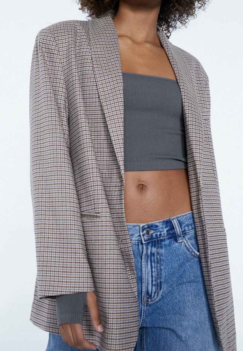 Femme portant un blazer oversized à carreaux, un top bandeau gris et un jean bleu taille haute, montrée du cou aux hanches sur fond blanc.