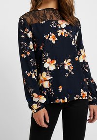 Blusa floreale in blu navy scuro con maniche lunghe, caratterizzata da un corpetto in pizzo, vivaci motivi floreali arancioni e gialli, e polsini elasticizzati.