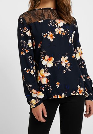 Blusa floreale in blu navy scuro con maniche lunghe, caratterizzata da un corpetto in pizzo, vivaci motivi floreali arancioni e gialli, e polsini elasticizzati.
