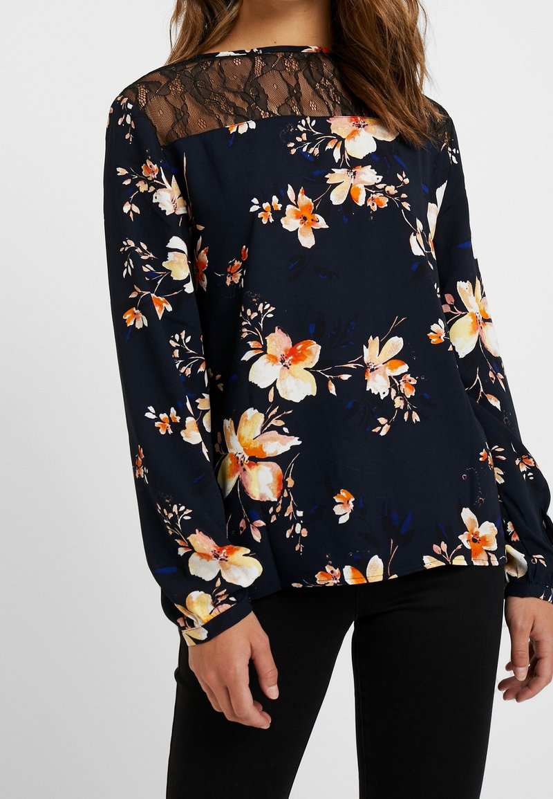 Blusa floreale in blu navy scuro con maniche lunghe, caratterizzata da un corpetto in pizzo, vivaci motivi floreali arancioni e gialli, e polsini elasticizzati.