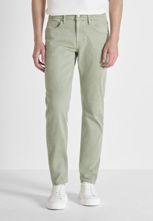 Man met lichtgroene slim-fit jeans en witte sneakers, staand tegen een effen lichte achtergrond.