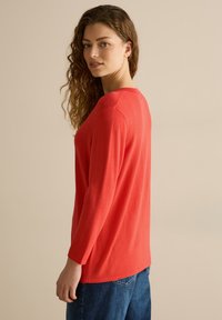 Rotes langärmeliges Oberteil mit Rundhalsausschnitt, weichem Material und lockerer Passform. Der Rücken ist etwas länger und wird mit lässigen blauen Jeans kombiniert.
