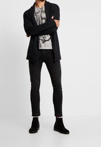 Černý cardigan přes šedé tričko s grafikou, spárovaný s úzkými černými džíny a černými semišovými kotníkovými botami. Žádné význačné vzory nebo textury.