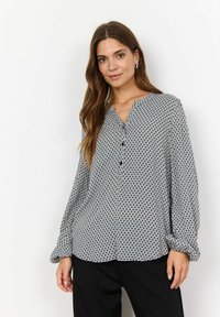 Blouse à motifs en tissu léger, présentant un design géométrique noir et blanc, avec une fermeture à bouton sur le devant et des manches longues avec des poignets élastiques.