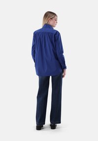 Oversized blue shirt with a rear yoke detail, buttoned cuffs, and a smooth texture, paired with dark denim wide-leg pants.