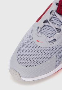 Zapatilla deportiva gris con parte superior de malla, suela de goma, acentos rojos y logo de Nike. Cuenta con cordones y un cuello acolchado para mayor comodidad.