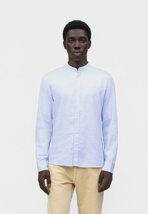 Νεαρός άνδρας με σκουρόχρωμο δέρμα, φορέοντας ένα ανοιχτό μπλε πουκάμισο με κουμπιά μακρυμάνικο και μπεζ παντελόνια, στέκεται μπροστά σε ένα απλό λευκό φόντο.