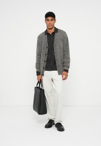 Grå strikket cardigan, sort polo shirt, hvide bukser, sorte sko. Holder en sort tote bag. Ensfarvet baggrund.