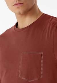 Bordeauxfarbenes T-Shirt mit kurzen Ärmeln und Rundhals, ausgestattet mit einer dezenten Tasche und gestreiftem Akzent. Weiche, glatte Textur aus Baumwollmischung.