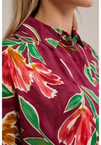 Bluză florală cu un fundal vișiniu și modele mari de flori roșii, roz și verzi. Design cu guler și material moale, texturat.