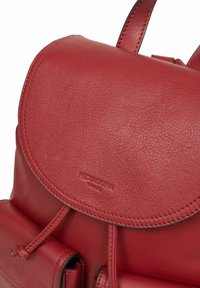 Rode leren rugzak met een afgeronde flap, een gestructureerde oppervlakte, verstelbare straps en een logo dat op de voorkant is gedrukt.