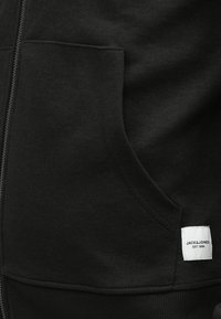 Fekete cipzáras kapucnis pulóver texturált anyagból, kenguruzsebbel és fehér márkacímkével az alján.