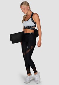 Leggings deportivos negros con paneles de malla y logo de la marca en blanco, combinados con un sujetador deportivo gris con una banda de logo negra. Llevando una esterilla de yoga.