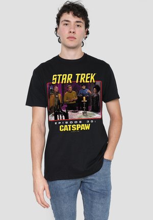 Junger Mann in schwarzem T-Shirt mit Grafik aus der Star Trek Folge "Catspaw" und blauen Jeans, vor einem einfarbigen hellgrauen Hintergrund stehend.