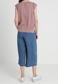 Blusa rossa a fantasia con maniche corte a balze e dettaglio a occhiello sulla schiena; abbinata a pantaloni culotte di jeans blu a gamba larga e sneakers bianche.