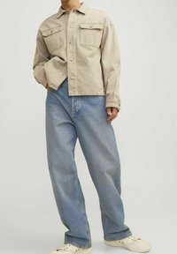 Beige denimjacka med två bröstfickor, ihop med ljusblå baggy jeans och krämfärgade sneakers. Tyget har en mjuk yta och en avslappnad passform.