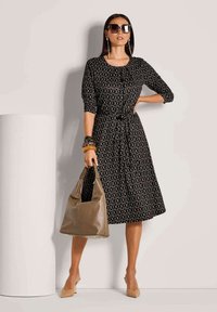 Frau in einem knielangen schwarzen gemusterten Kleid, mit großen Sonnenbrillen, beigen Absätzen und Armbändern, die eine taupefarbene Lederhandtasche trägt.