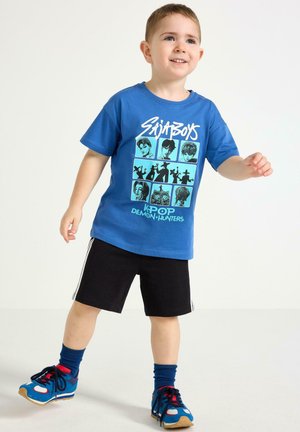 Ung dreng smilende, iført en blå T-shirt med grafik "SK8BOYS KPOP DEMON HUNTERS", sorte shorts, blå sokker og blå sneakers.