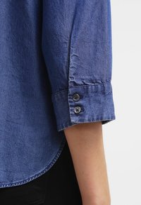 Chemise en denim bleu avec des manches repliées et deux boutons noirs, présentant une texture lisse et un éclat subtil. L'ourlet est arrondi.