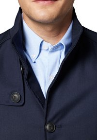 Marynarka w kolorze granatowym z wysokim kołnierzem i dwoma czarnymi guzikami, nałożona na jasnoniebieską koszulę w jodełkę z białymi guzikami.