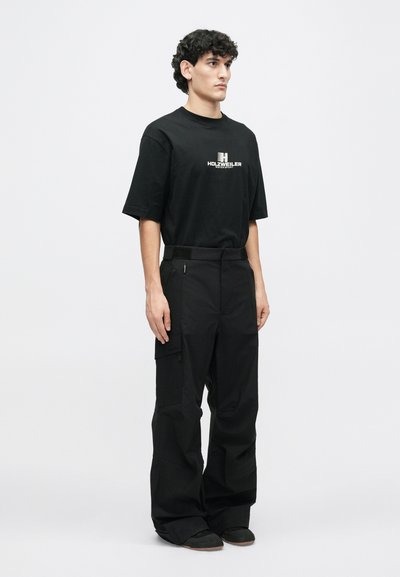 Juodas trumpas rankovės marškinėliai su baltu logotipo, derinami su juodomis laisvomis cargo kelnėmis su šoninėmis kišenėmis ir velcro akcentais.