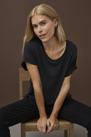 Fiatal nő hosszú szőke hajjal, fekete ingben és nadrágban, egy fából készült széken ül, barna háttér előtt.