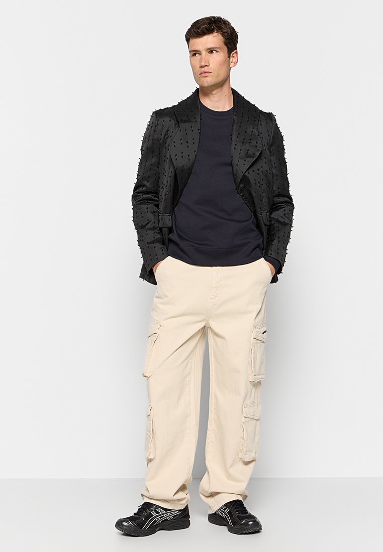 Veste noire texturée au design ajusté, associée à un T-shirt bleu marine et un pantalon cargo beige. Comprend des poches et une silhouette décontractée.