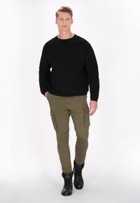 Černá mikina, olivové cargo kalhoty s bočními kapsami a černé boty. Model stojí čelem k fotoaparátu na bílém pozadí.