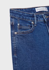 Jeans in denim blu con un classico design a cinque tasche, cuciture in rame e un'etichetta sul cinturino con il marchio. Texture liscia senza un motivo visibile.