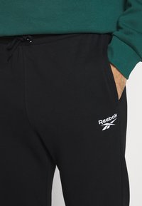 Pantalon de survêtement noir avec cordon de serrage, présentant un logo Reebok blanc sur la cuisse. Fabriqué en tissu doux avec une texture lisse.
