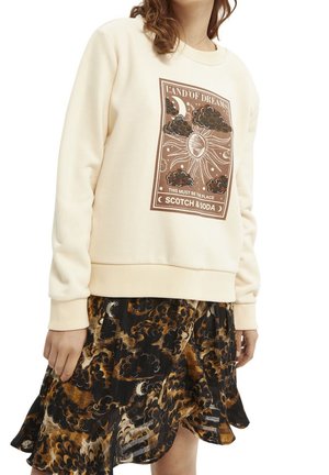 Femme portant un sweat crème avec un motif soleil, lune et nuages ainsi qu’un texte, associé à une jupe noire et marron à motif.