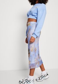 Mujer vestida con un sudadera corta azul claro, falda midi con estampado de querubines, calcetines blancos y zapatillas deportivas blancas, de pie contra un fondo liso.