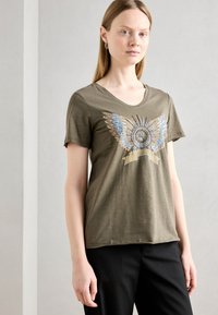 Olijfgroene T-shirt met een V-hals, voorzien van decoratieve vleugeldetails in blauw en goud, gecombineerd met zwarte broek.