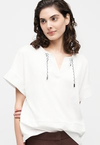 Γυναίκα με σγουρά μαλλιά μέχρι τους ώμους, που φοράει λευκή, φαρδιά μπλούζα με κοντά μανίκια και μαύρα και λευκά κορδόνια στον γιακά.