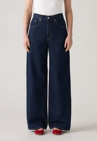 Jeans din denim albastru închis, cu croială largă, talie înaltă, design clasic cu cinci buzunare și detalii cusute galbene. Asortate cu pantofi roșii.