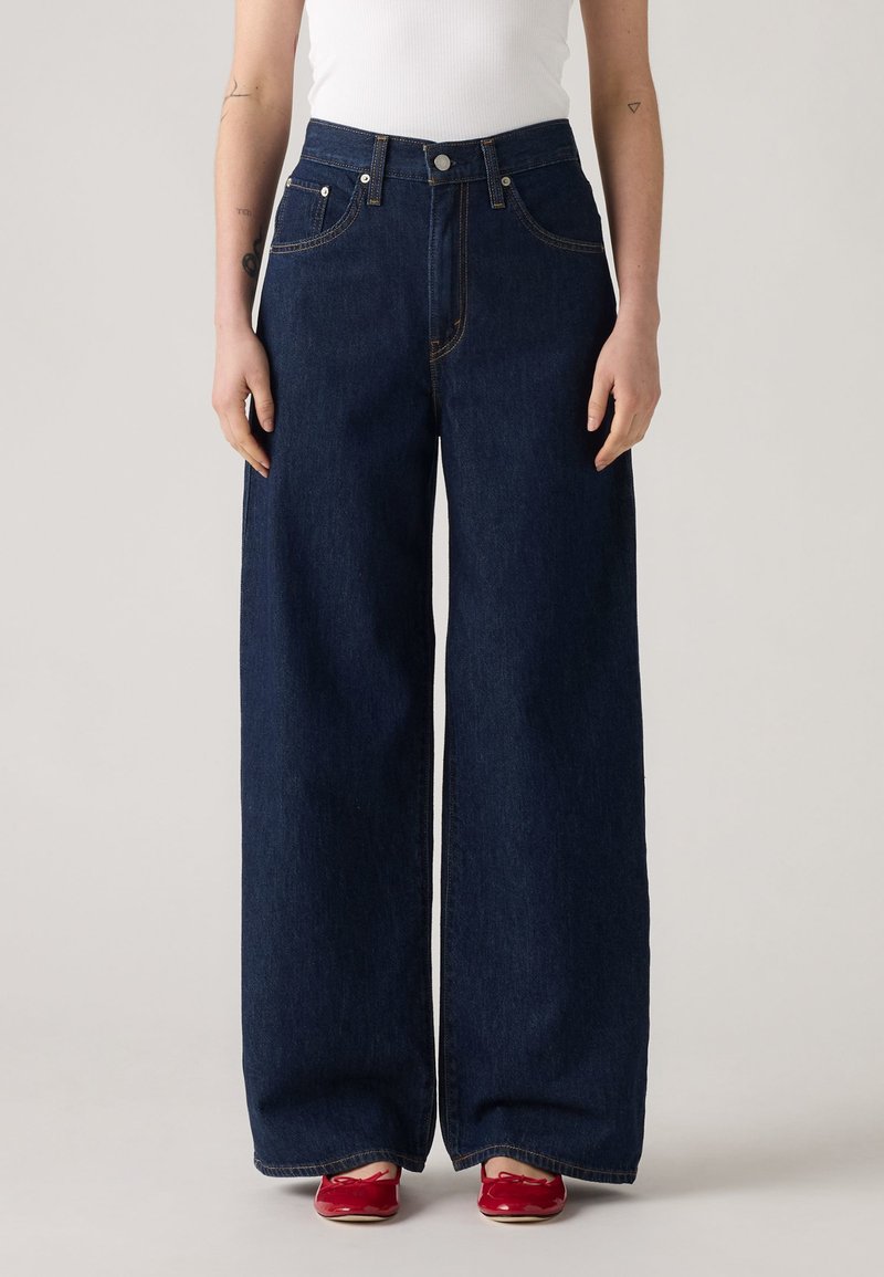 Jeans din denim albastru închis, cu croială largă, talie înaltă, design clasic cu cinci buzunare și detalii cusute galbene. Asortate cu pantofi roșii.