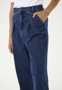 Tummansinisiä korkeavyötäröisiä farkkuhousuja, joissa on sivutaskut, laskosyksityiskohdat ja sileä rakenne, suunniteltu rentoa istuvuutta varten.