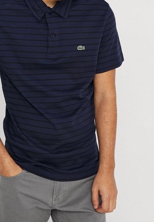 Mann trägt ein dunkelblau gestreiftes Poloshirt mit grünem Krokodil-Logo und graue Hose, die Hände locker an den Seiten.