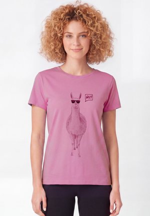 Frau mit lockigem Haar, die ein pinkes T-Shirt mit einem Lama in Sonnenbrille und einer Sprechblase mit dem Text "Hi!" trägt