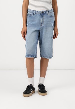 Shorts en denim bleu clair, à coupe ample, avec des bords effilochés et un aspect usé. Portés avec des chaussettes blanches et des baskets noires.