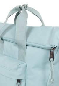 Ljusblå väska i ett texturerat tyg, med en överfäll, två robusta handtag och en dragkedjeficka med en dekorativ dragare.