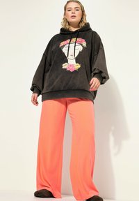 Přesně padnoucí černá mikina s kapucí zdobená květinovým motivem, kombinovaná s korálovými kalhotami s vysokým pasem a širokými nohavicemi vyrobenými z jemné hladké látky. Viditelné ploché boty.