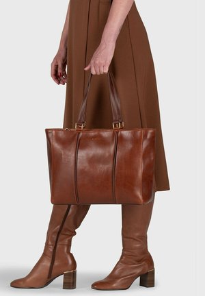 Femme portant des bottes marron et une robe, tenant un grand sac fourre-tout en cuir marron avec deux poignées, sur un fond uni.